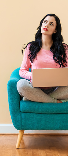 A woman in a pink shirt sitting cross-legged on a chair with a laptop in her lap thinking about credit